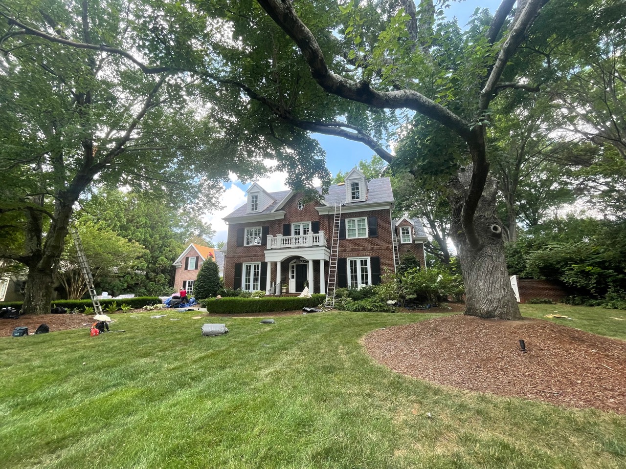 Front view of an active roof installation on a brick colonial residence surrounded by mature trees and landscaping